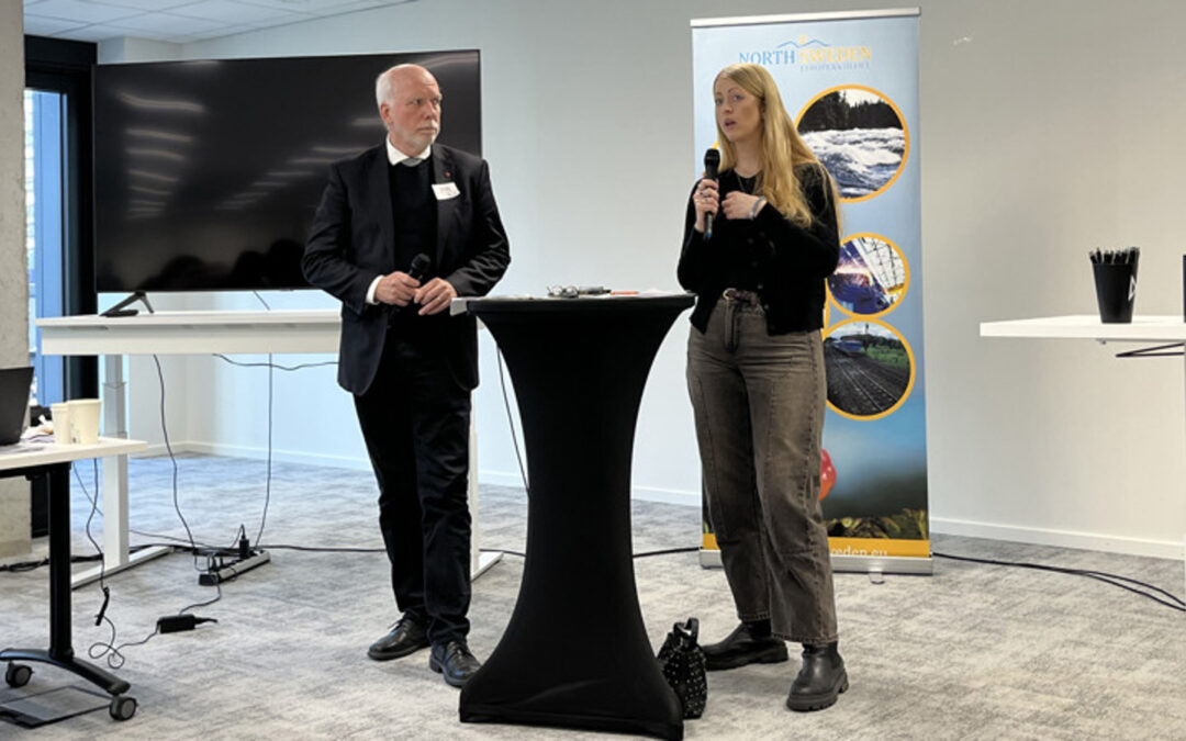 Två personer står vid ett ståbord. En av dem pratar i en mikrofon. Den andra lyssnar. I bakgrunden syns en storbildsskärm och en roll up från North Sweden European Office.