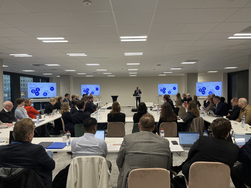 Ett 60-tal deltagare lyssnar på en talare som står längst fram i möteslokalen. På sidorna om talaren syns fyra storbildsskärmar som visar samma presentation så att den ska synas oavsett var i lokalen åhörarna sitter.