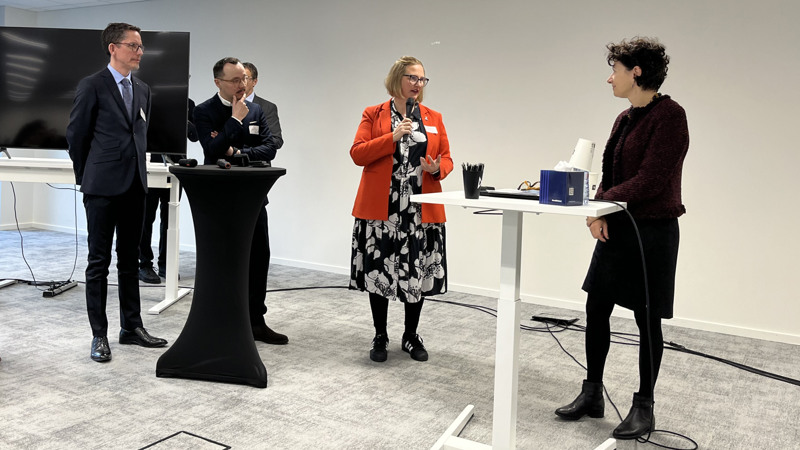 En person med röd kavaj och svart klänning med vita blommor på står mitt i bilden och pratar i mikrofon. Hon riktar blicken mot en person med mörk tröja och mörk kjol som står till höger i bild. Till vänster om talaren står två personer i mörka kostymer och lyssnar.